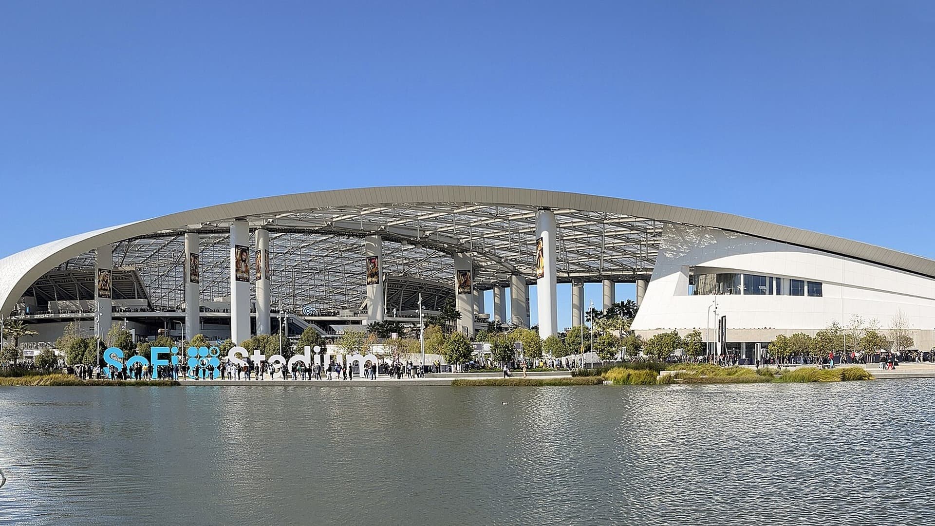Wide exterior view of SoFi Stadium in Inglewood in April 2023, with the SoFi Stadium sign beside the water.