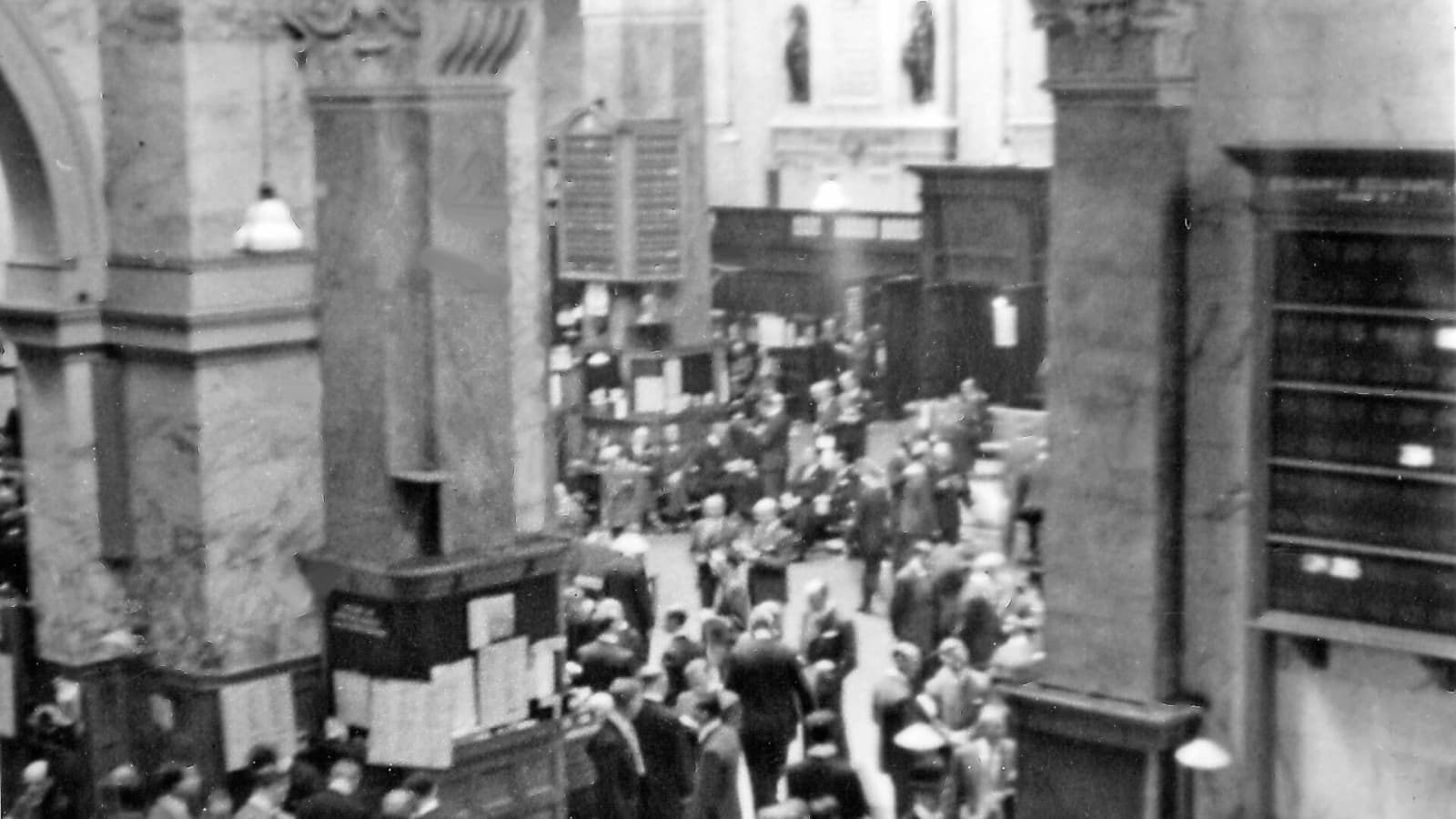 A historic black-and-white photograph of the London Stock Exchange trading floor filled with market participants