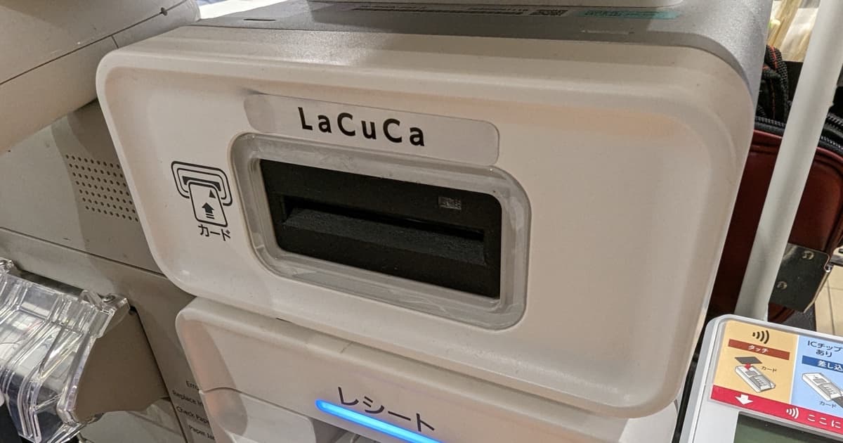 Un terminal de paiement blanc en magasin, avec une fente pour carte et un symbole sans contact, près d'une caisse de supermarché.