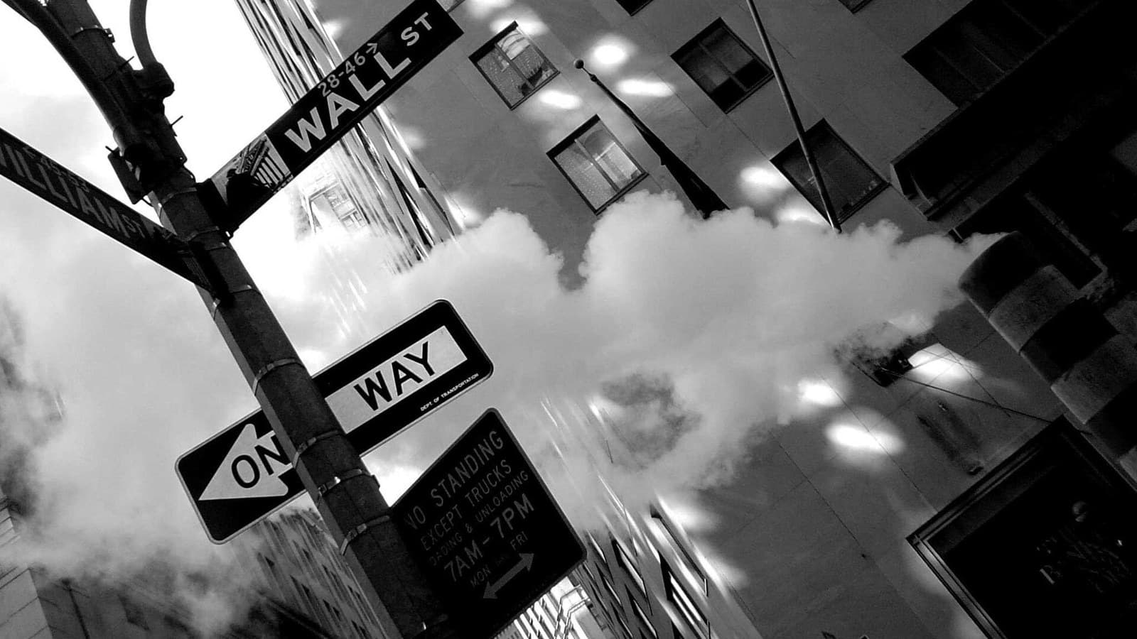 Black-and-white view down Wall Street between tall Financial District buildings above a roadworks steam stack