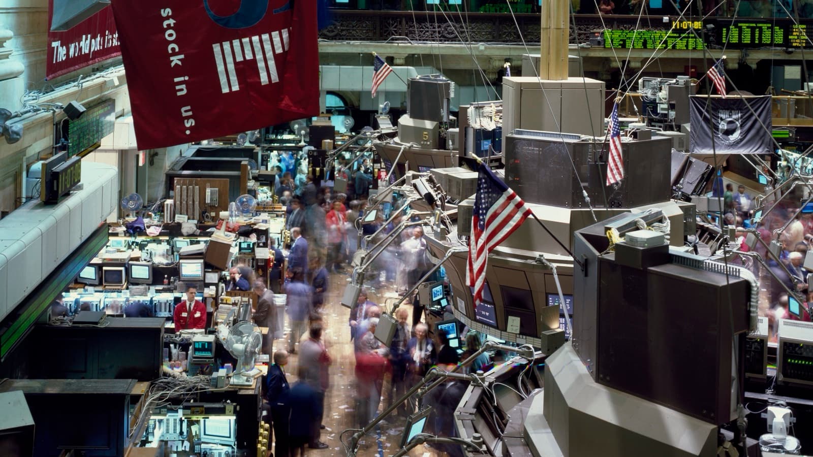 A busy New York Stock Exchange floor with moving traders, workstation posts, quote boards and American flags
