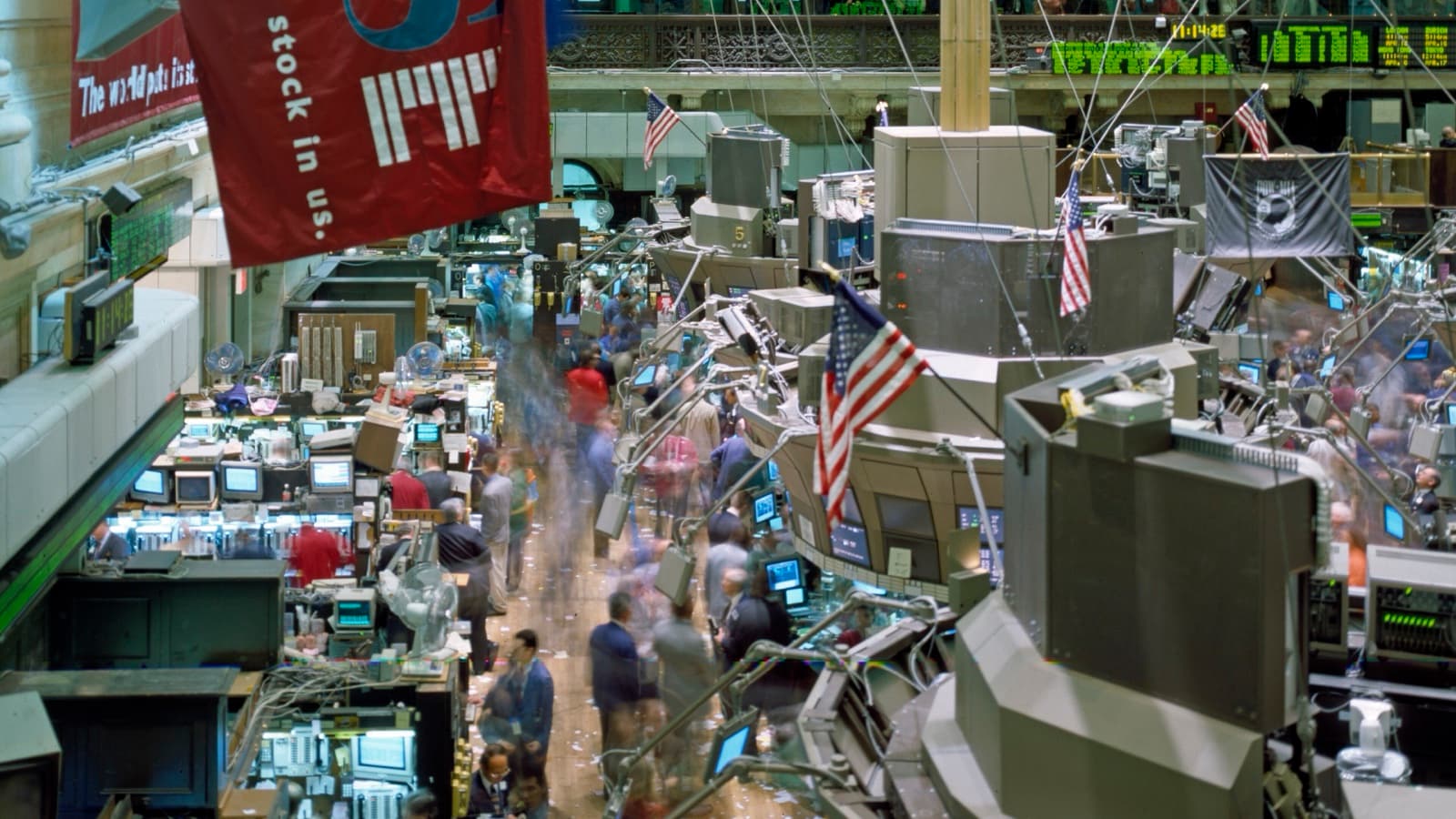 A busy New York Stock Exchange floor with moving traders, workstation posts, quote boards, American flags and a large red market banner