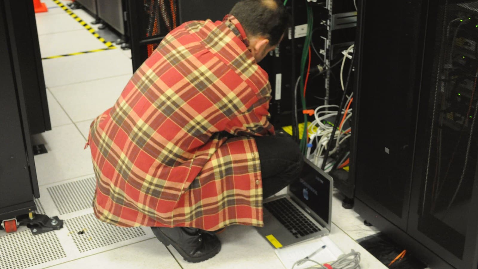A technician crouches with a laptop beside an open server rack in a data centre