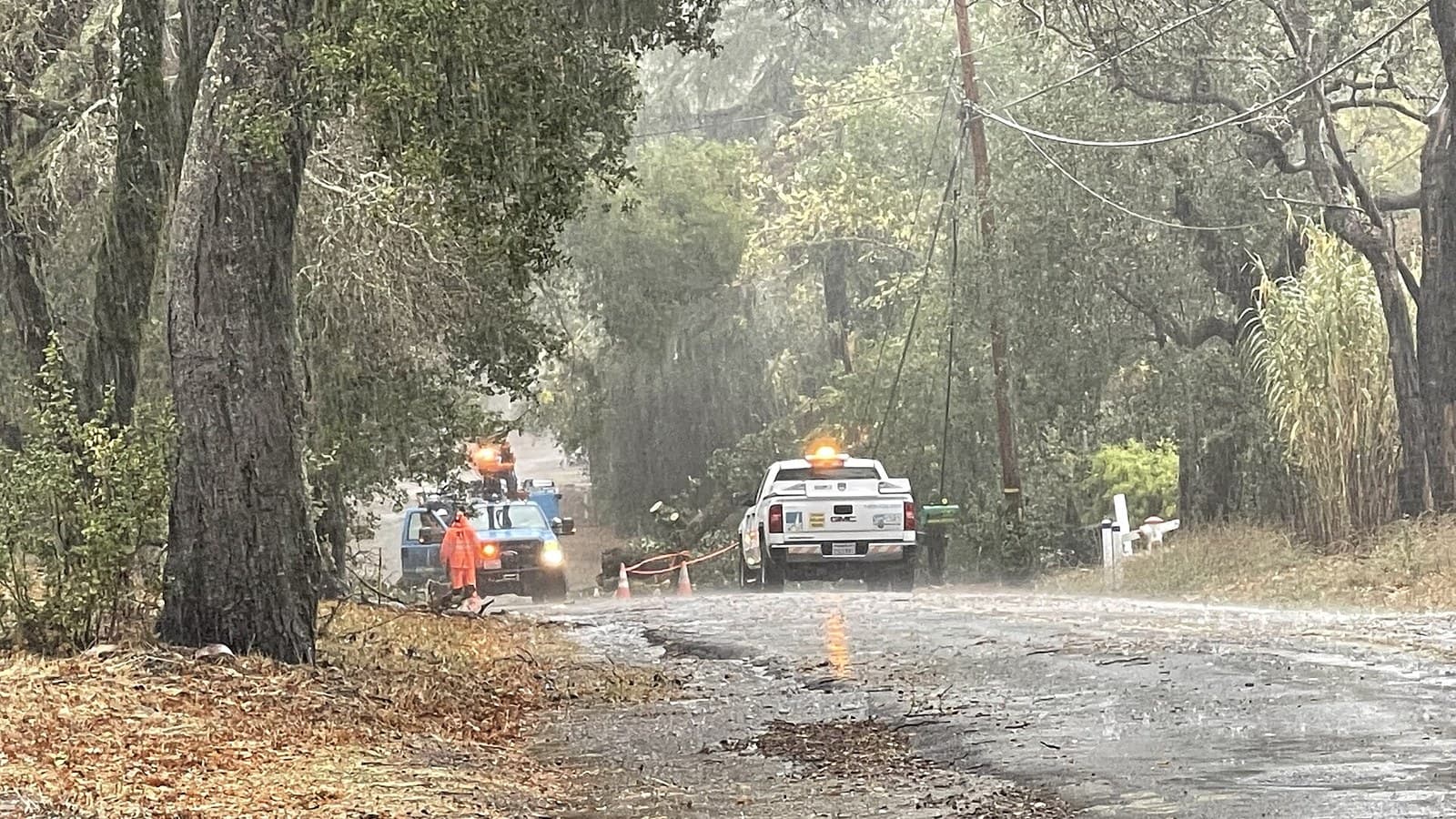 Two PG&E utility trucks and a high-visibility worker respond to downed lines on a rain-soaked California road