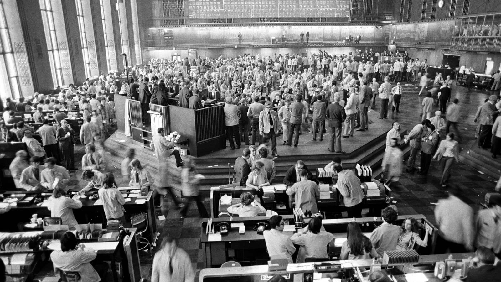 Black-and-white photograph of a crowded commodities trading floor beneath a large quote board