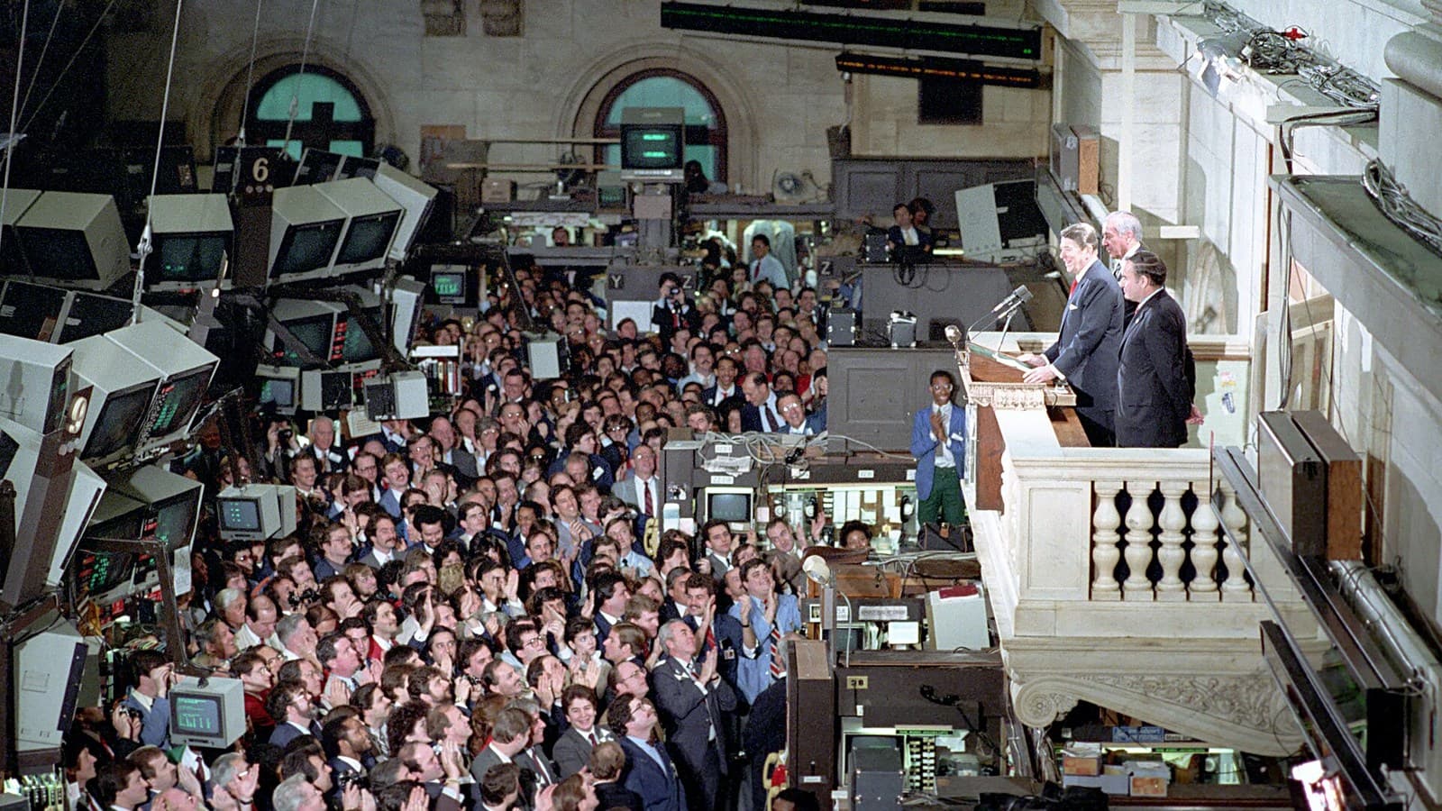 Archival view of the crowded New York Stock Exchange floor in 1985, with CRT screens and a speaker on a balcony.