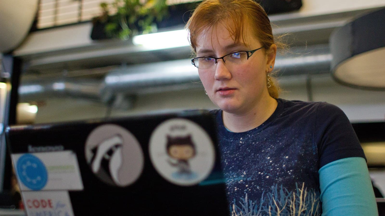 A programmer with glasses works behind a stickered laptop at a San Francisco hackathon