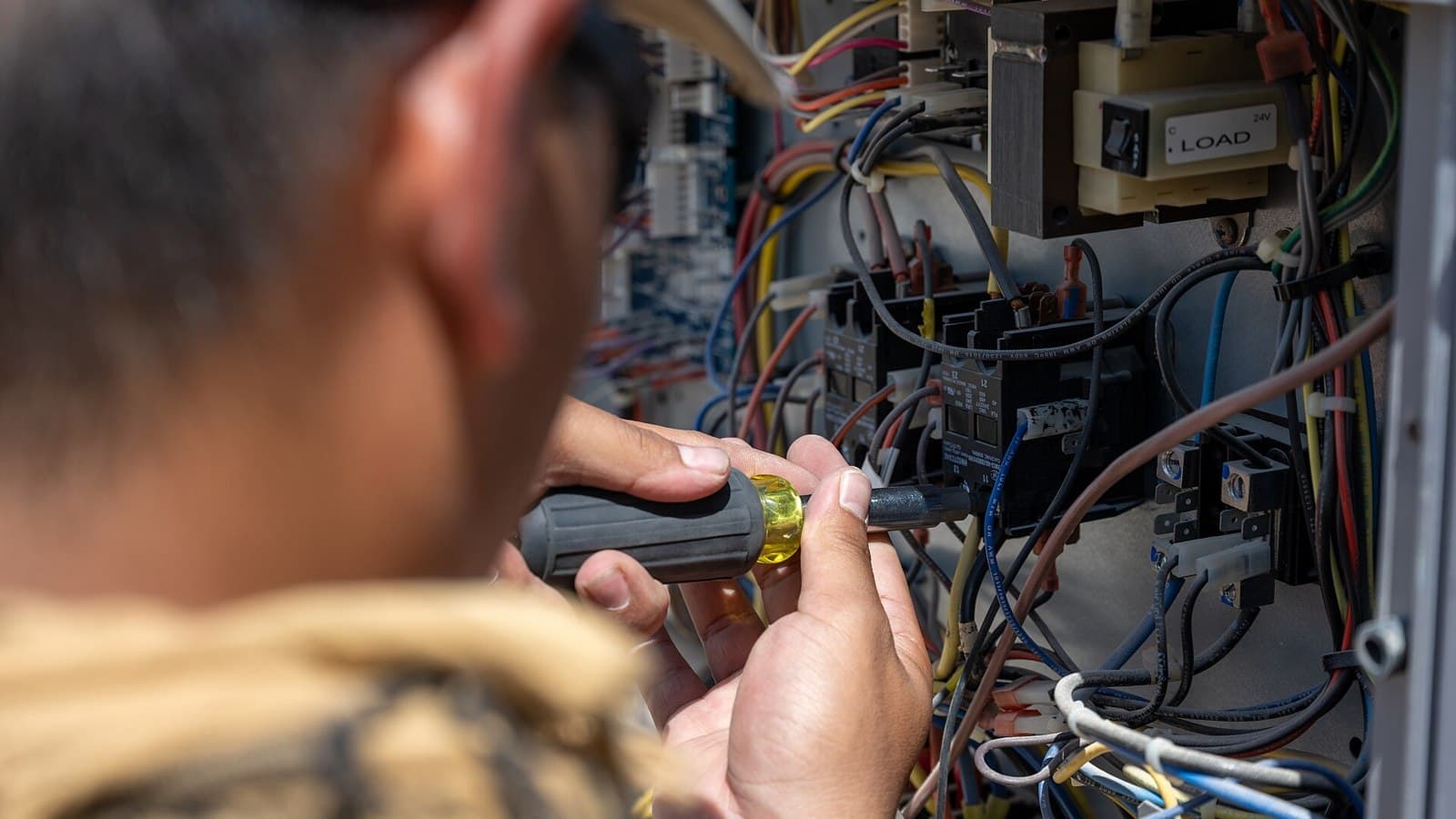 Technician using a screwdriver inside an open electrical control panel with multicolored wiring.