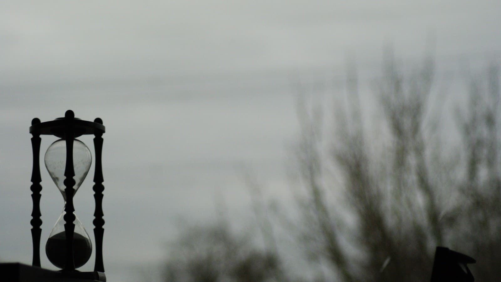 Dark-framed hourglass against a softly blurred sky and bare trees